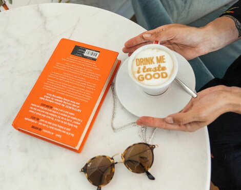 Eine Person genießt eine Tasse Kaffee mit Latte Art und ein Buch auf einem runden Tisch, möglicherweise in der Lounge des Strandhotels Ahlbeck, Usedom.