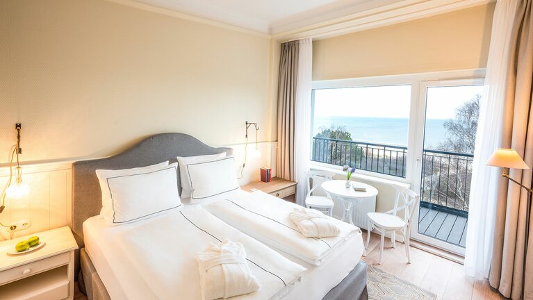 Elegantes Deluxe Doppelzimmer im Strandhotel Ahlbeck mit einem komfortablen Doppelbett, einem Balkon mit Meerblick und Bademänteln auf dem Bett.